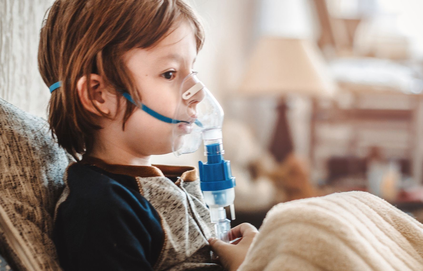 Child using a nebulizer mask while sitting on a sofa, holding a blanket.